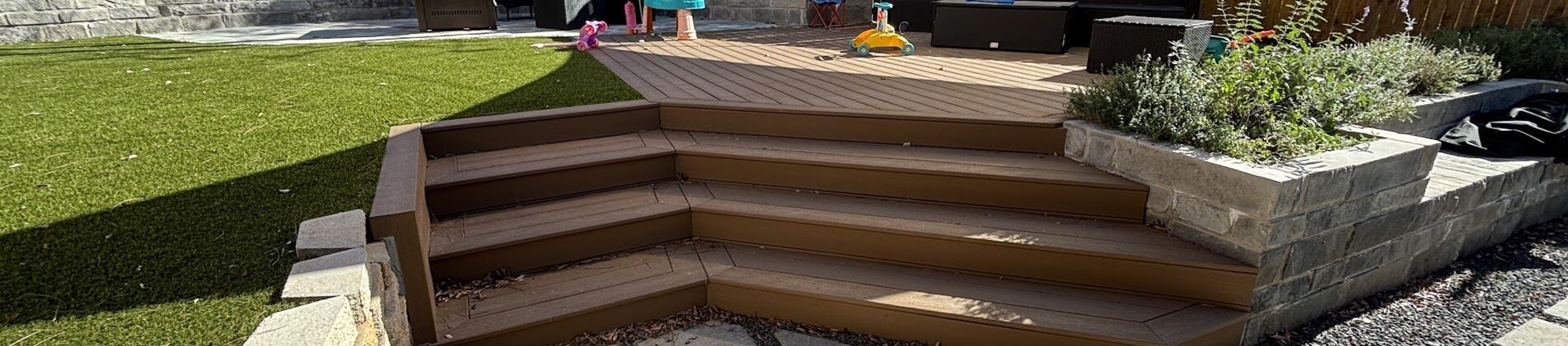 Austin backyard transformation with artificial turf, a composite deck with wraparound steps, a stone block planter, and concrete pavers in gravel