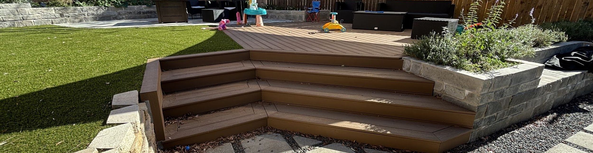 Austin backyard transformation with artificial turf, a composite deck with wraparound steps, a stone block planter, and concrete pavers in gravel