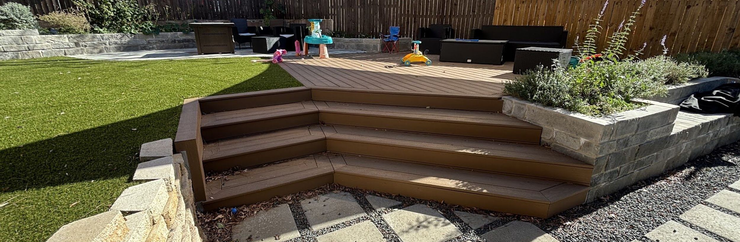 Austin backyard transformation with artificial turf, a composite deck with wraparound steps, a stone block planter, and concrete pavers in gravel