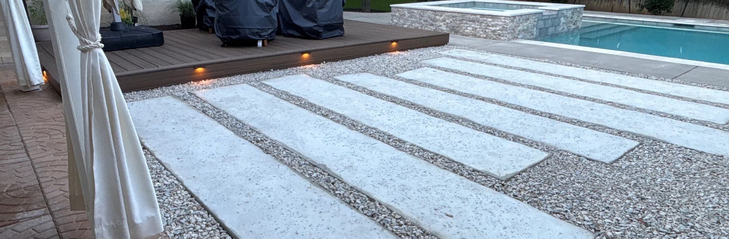 Backyard with a lit composite deck, large concrete pavers set in gravel, and a pool and spa in Austin