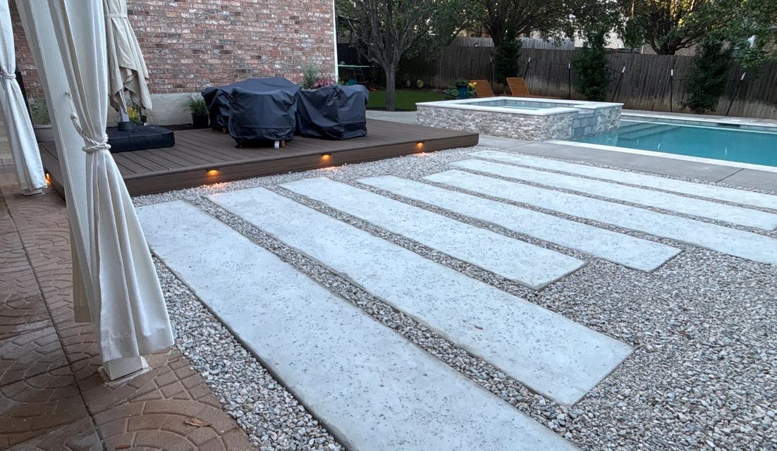 Backyard landscape design in Austin with a lit composite deck, large concrete pavers in gravel, and a pool and spa