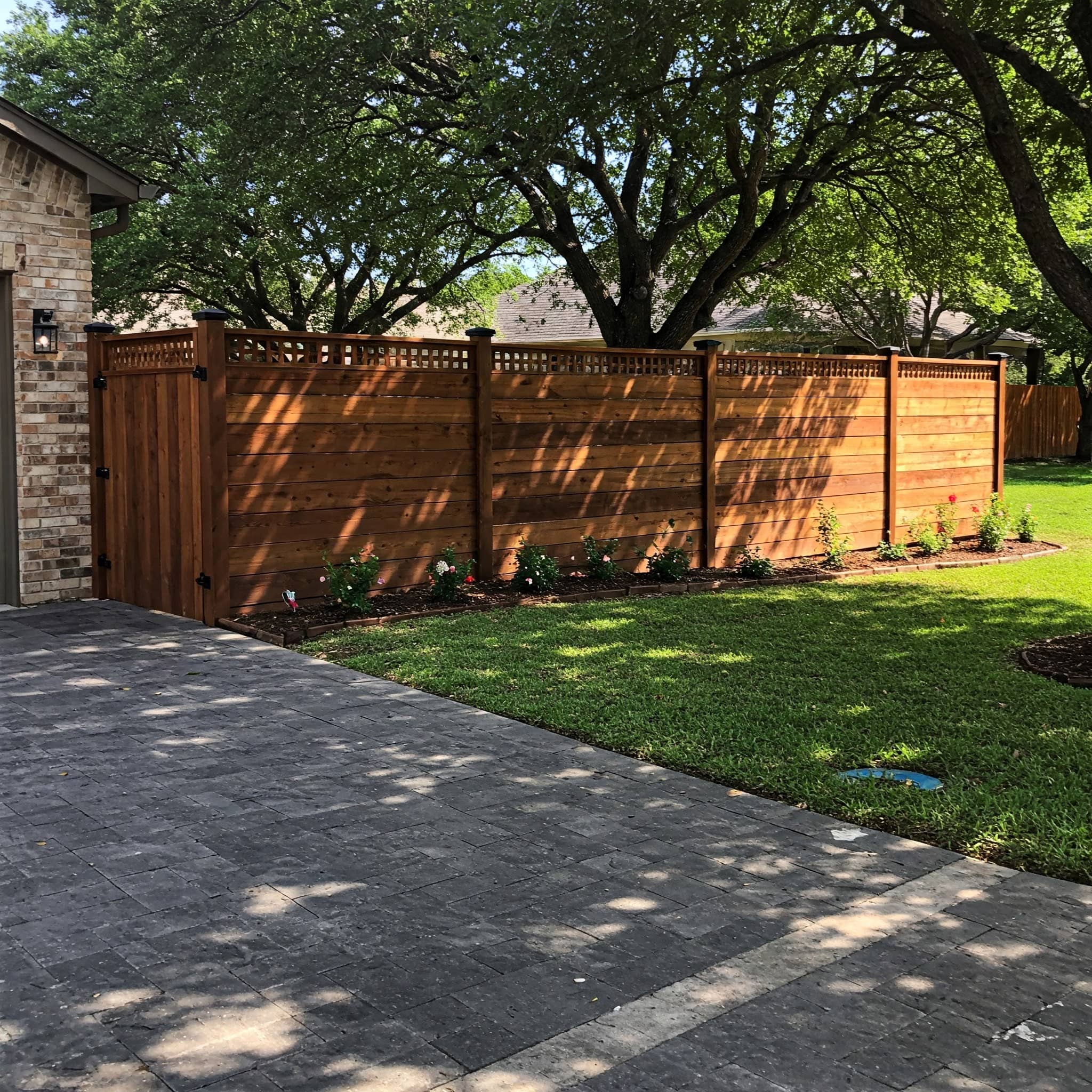 Stained horizontal cedar privacy fence with a lattice top and matching gate beside a paver driveway, shaded by mature live oaks