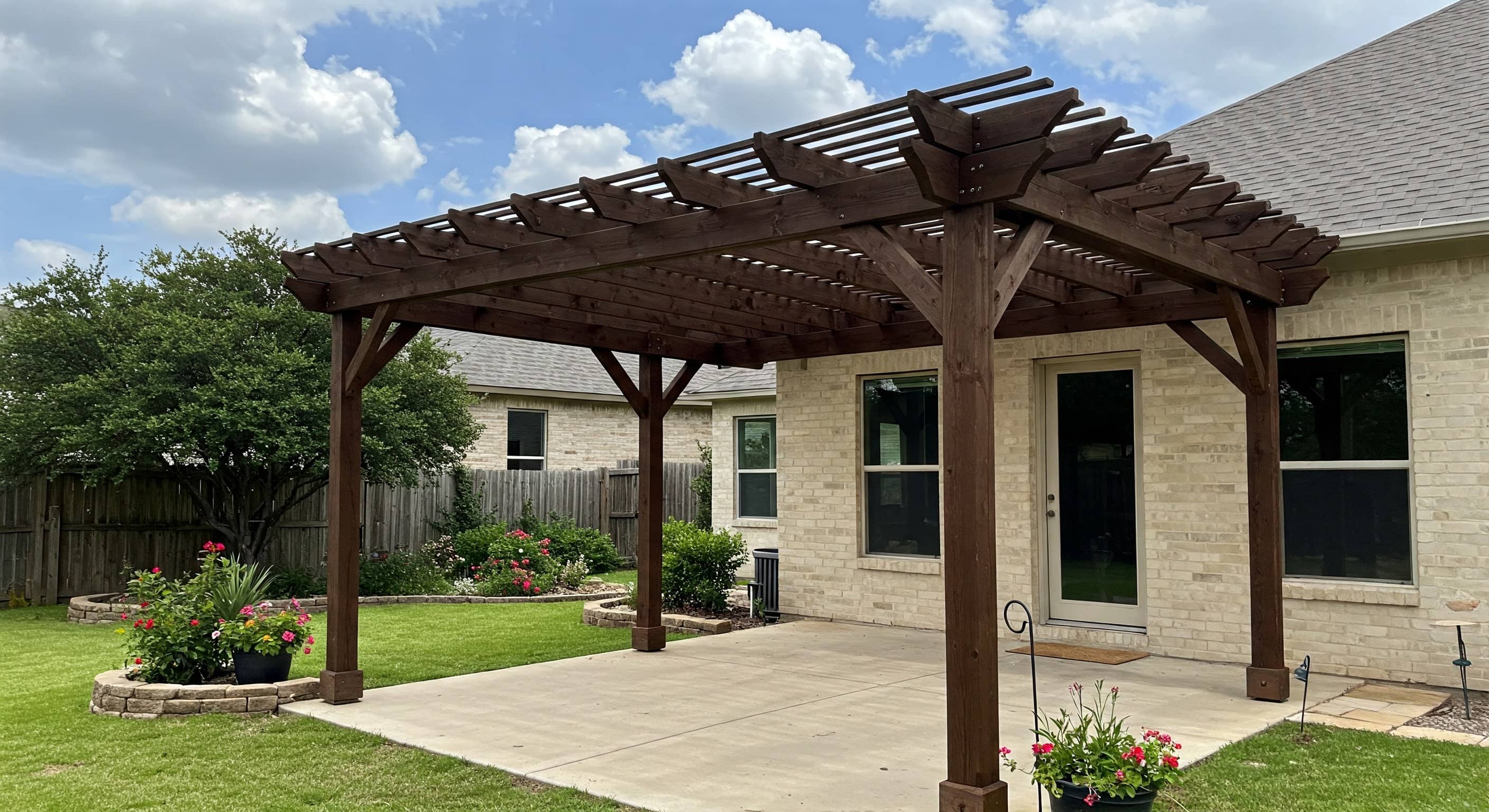 Wood pergola over a concrete patio beside a brick home and raised garden beds